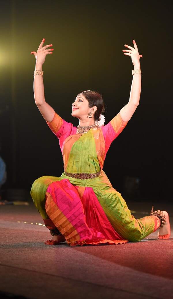 Dakshina in a colourful Bharatanatyam costume, both arms lifted during a stage performance